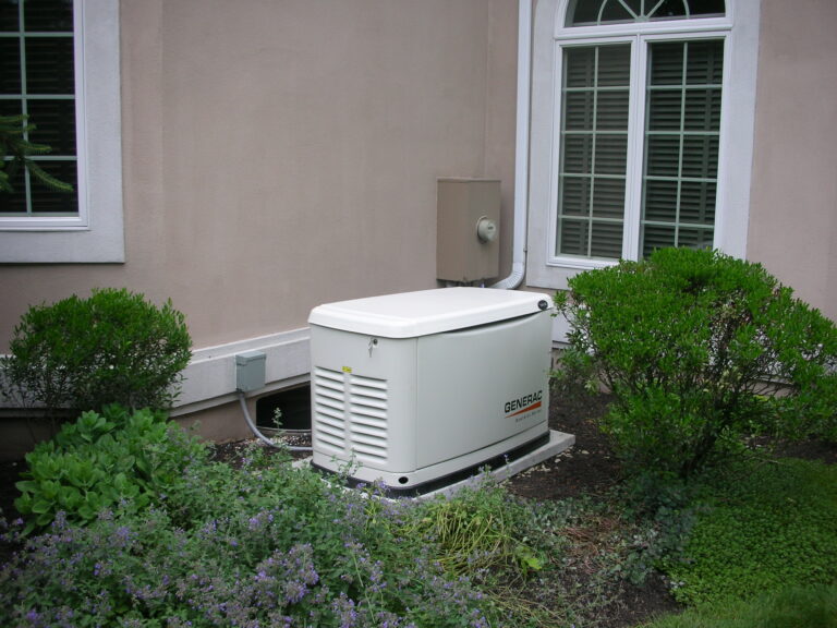 A standby generator, professionally installed by Magic Electric, Plumbing, Heating & Air, sits outside a customer’s home near a window and is surrounded by well-maintained shrubs and plants.