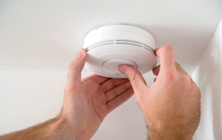 A professional Magic Electric, Plumbing, Heating & Air technician carefully installing a white smoke detector on a ceiling.